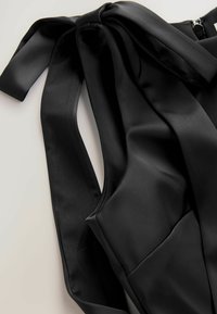Top din satin negru cu o fundiță la umăr. Are o textură netedă și un sistem de închidere cu fermoar. Materialul este drapat și lucios.