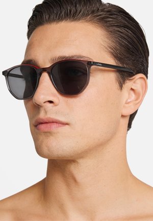 Occhiali da sole marroni con lenti scure, forma angolare e una montatura bicolore con una fodera interna rossa e un bordo esterno nero.