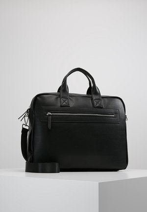 Malette en cuir noir avec double poignée, poche avant zippée et bandoulière amovible, posée sur un socle blanc sur fond gris.