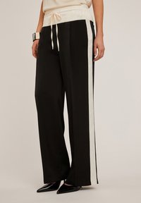 Svarta vida byxor med en krämfärgad elastisk midja, som har dragsko och en krämfärgad sidragge, tillverkade av ett mjukt, lätt tyg.