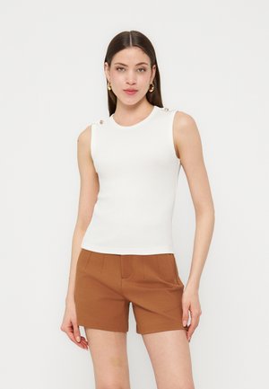 Kvinde med langt mørkt hår iført hvid ærmeløs top med skulderknapper og brune shorts med høj talje, står foran en ensfarvet baggrund.