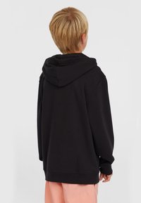 Sudadera negra con capucha con cordones, de corte holgado y puños acanalados. La tela parece suave, llevada sobre pantalones rosa claro. Vista trasera.