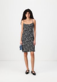 Rochie neagră fără mâneci, cu un model abstract alb, tăiat la genunchi și cu bretele subțiri. Modelul ține o geantă albastră și poartă pantofi negri.
