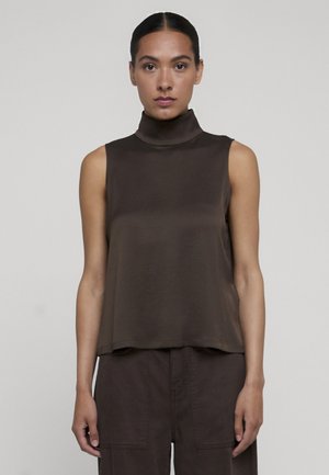 Frau mit dunklen Haaren zu einem eleganten Dutt gebunden, trägt ein ärmelloses dunkelbraunes Rollkragentop und passende dunkelbraune Hose vor neutralem Hintergrund.