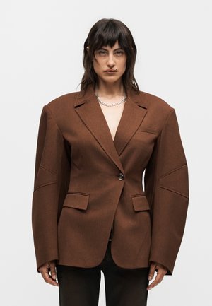 Γυναίκα με σκούρα μαλλιά που φοράει υπερμεγέθη καφέ σακάκι με υπερβολικά μαξιλαρωμένους ώμους και μαύρο παντελόνι, κοιτάζοντας μπροστά σε απλό φόντο.