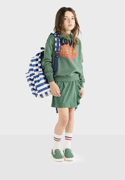 Felpa verde con una grafica rossa e una gonna verde a volant. La modella porta uno zaino a strisce blu e bianche e indossa delle scarpe slip-on verdi.