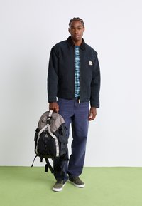 Black jacket with a brown collar and front pocket, striped blue and white shirt, dark pants, and a gray and black backpack with straps.