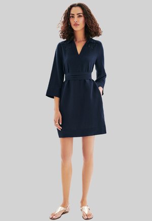 Femme aux cheveux bouclés portant une robe bleu marine jusqu'aux genoux avec des manches 3/4 et une ceinture, chaussée de sandales dorées, se tenant devant un fond gris clair.