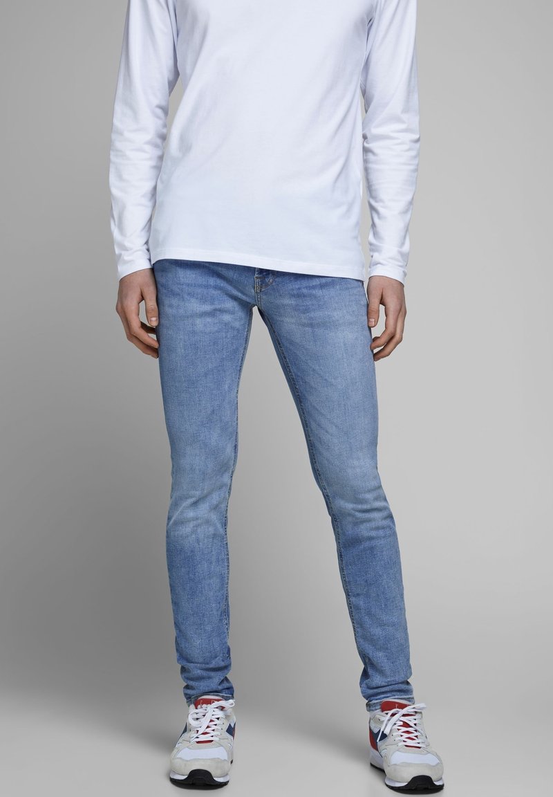 Jeans ajustados de mezclilla en azul claro, con un diseño clásico de cinco bolsillos, sutiles desgastes y cintura de tiro medio. Combinados con una camiseta blanca de manga larga y zapatillas.