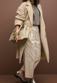 Femme portant un long manteau beige, un pull gris, un pantalon beige taille haute, des mocassins métalliques, tenant un grand sac à main beige.