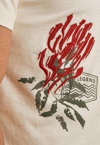 T-shirt couleur crème avec un design de plante brodé rouge et des feuilles vertes, accompagné d'un logo rectangulaire en arrière-plan.