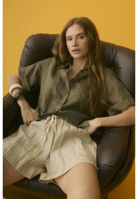 Femme aux longs cheveux bruns assise détendue dans un fauteuil en cuir sombre, portant une chemise olive, un short beige à rayures et des bracelets épais sur fond jaune.