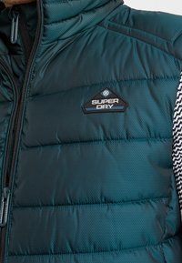 Blågrön pufferväst med quiltad textur, svarta accenter och en Superdry-logotyp. Har en dragkedja fram och kontrasterande mönstrade sidopaneler.