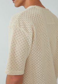 Beige gebreid sweater met een losse, ademende textuur met een ruitpatroon en korte mouwen. Ronde halslijn en subtiele geribbelde accenten.