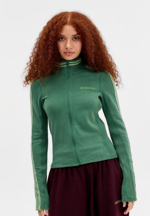 Žena s kučeravými červenými vlasmi oblečená v zelenú bundu na zips s pásikavými rukávmi a bordovými nohavicami, stojaca pred jednoduchým svetlým pozadím.