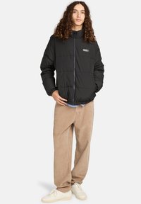 Ung person med langt krøllet hår iført en sort pufferjakke, beige fløjl bukser og hvide sneakers, stående med hænderne i lommerne.