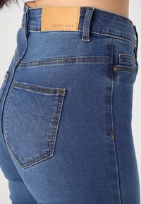 Jean en denim bleu taille haute avec un patch en cuir portant l'étiquette "NOISY MAY", doté d'un design classique à cinq poches et de coutures contrastantes.