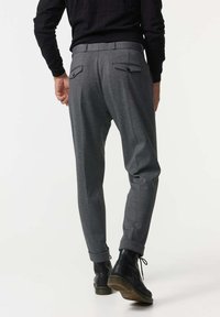 Graue schmal zulaufende Hose mit einem Bund und zwei Gesäßtaschen. Getragen mit schwarzen Stiefeletten. Glatte Stofftextur, minimalistisches Design.