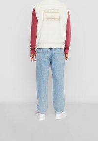 Biała kamizelka sherpa z naszywką z logo, bordowa koszula z długim rękawem, jasnoniebieskie luźne dżinsy i białe trampki. Swobodny strój widziany od tyłu.