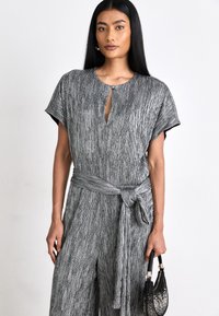 Grå tekstureret jumpsuit med korte ærmer, nøglehul-udskæring, matchende bælte i taljen og en dekorativ sort håndtaske.