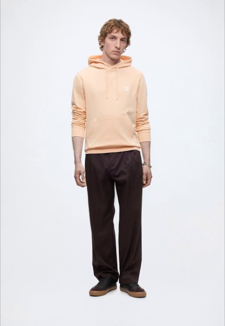 Jeune homme portant un sweat à capuche pêche avec poche avant, pantalon large foncé, baskets noires avec semelles en gomme, debout devant un fond clair uni.