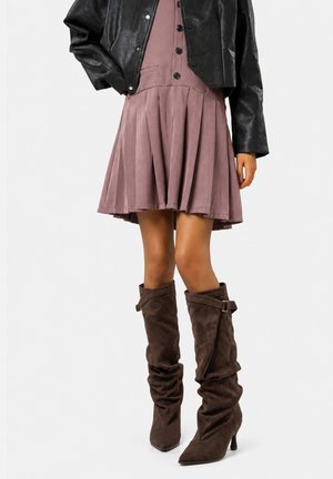 Mujer con vestido malva abotonado, chaqueta corta de cuero negra y botas marrones de ante holgadas hasta la rodilla con tacones.