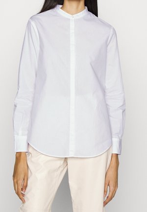 Mujer con una camisa blanca de manga larga con cuello mao y pantalones beige claro, de pie frente a un fondo liso.