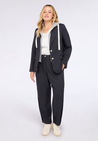 Kobieta w czarnym garniturze w prążki z białymi guzikami, szarym dresowym kapturkiem, białej bluzce i beżowych trampkach, stojąca na tle białego tła.