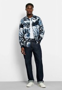 Blau-weiß gemusterte Jacke mit Landschaftsdesign, getragen über einem weißen T-Shirt. Kombiniert mit dunklen Jeans und weißen Sneakers.