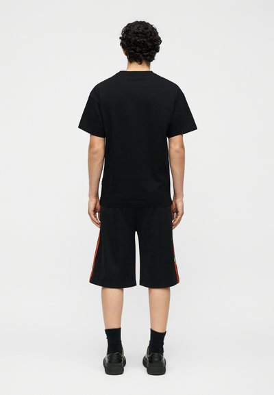 Άνδρας με σγουρά μαλλιά που φοράει απλό μαύρο μπλουζάκι, μαύρα σορτς μέχρι το γόνατο με κόκκινες ρίγες, μαύρες κάλτσες και μαύρα παπούτσια, γυρισμένος με την πλάτη.