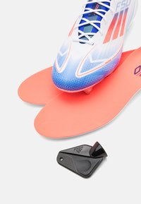 Hvit og blå fotballsko med røde detaljer, ledsaget av to rosa innersåler og et svart sportsaccessoire med teksturerte detaljer.
