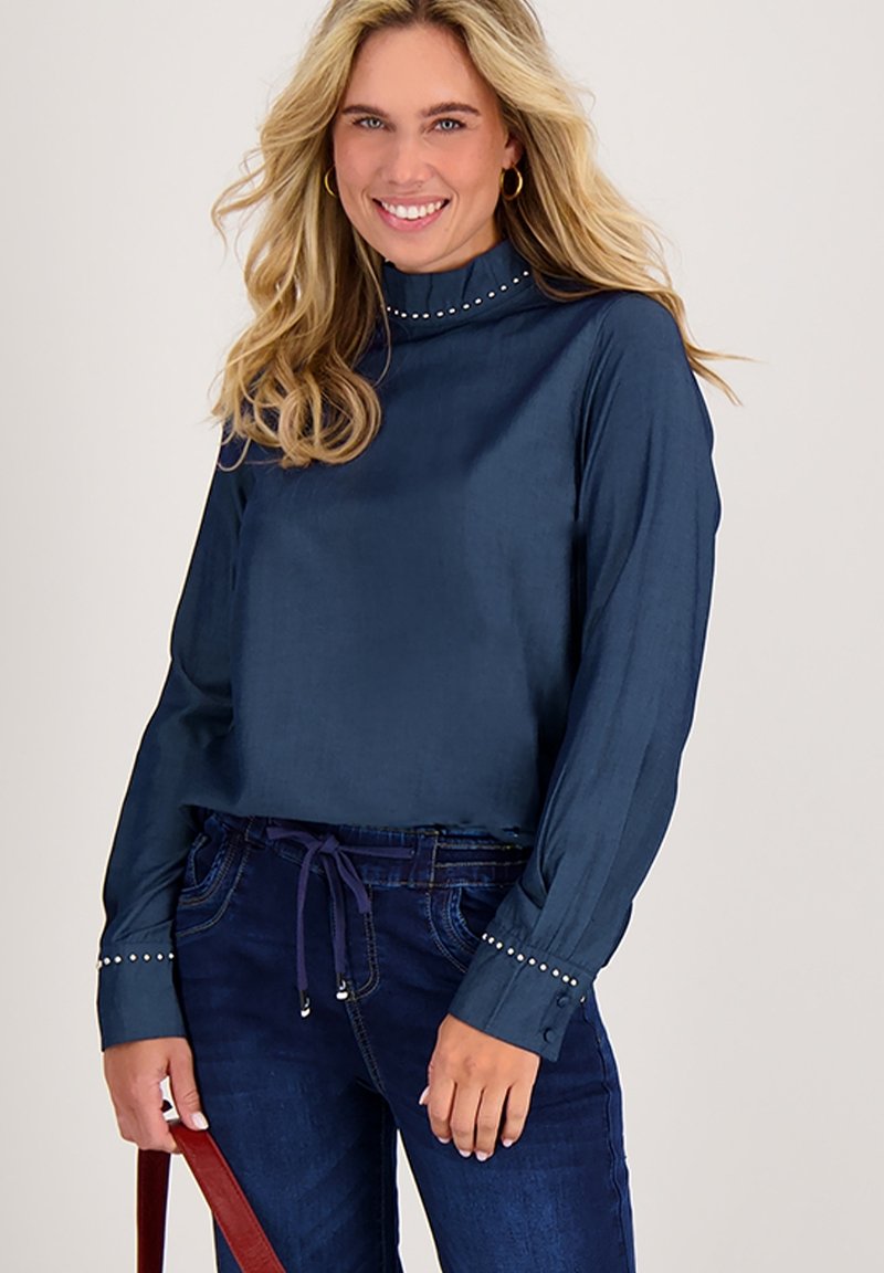 Donkerblauwe blouse met lange mouwen en hoge kraag, versierd met studs op de manchetten, gemaakt van een gladde, lichte stof. Gecombineerd met donkere jeans.