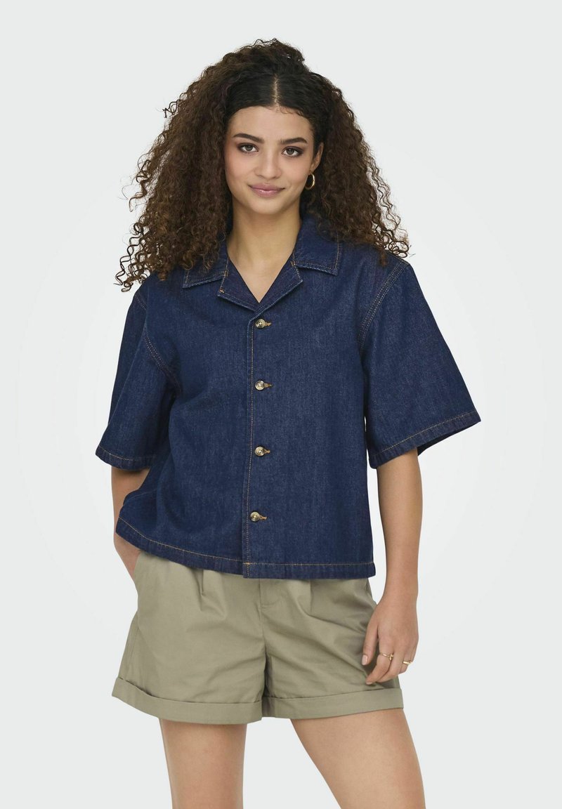 Camicia in denim blu scuro a maniche corte con colletto, caratterizzata da grandi bottoni e una vestibilità rilassata. Abbinata a pantaloni corti beige chiaro.