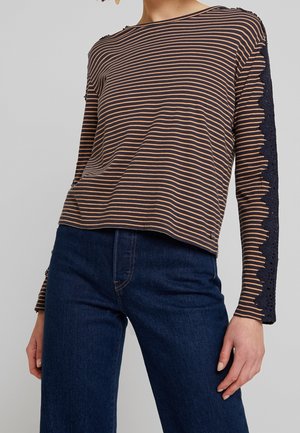 Kobieta ubrana w granatowo-beżowy prążkowany top z długim rękawem, wykończony granatową koronką na rękawach, oraz ciemnoniebieskie, wysokie spodnie jeansowe.