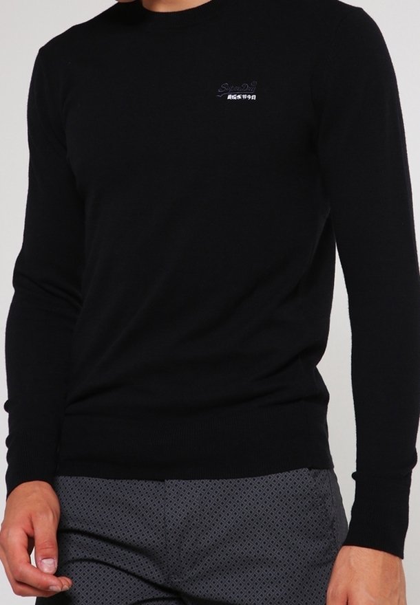 Personne portant un pull noir ajusté à manches longues avec un petit texte de logo blanc sur la poitrine et un pantalon gris à motifs.