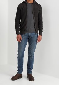 Czarny dzianinowy kardigan na zamek, noszony na szarym t-shircie, w połączeniu z niebieskimi dżinsami i brązowymi skórzanymi butami. Kardigan ma teksturowane detale.