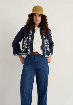 Frau mit lockigem Haar, die einen geflochtenen beigen Hut, eine marineblaue bestickte Jacke, eine weiße Bluse, einen braunen Gürtel und hoch taillierte blaue Jeans trägt.