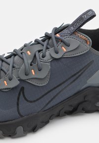 Nike-skor med övre del i grått tyg, strukturerade detaljer och en svart gummisula. Har orange snören och ett diagonalt logoband.
