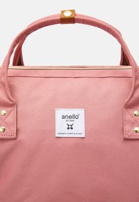 Sac fourre-tout en toile rose avec poignées doubles, accents en métal doré, et une étiquette blanche portant le nom de la marque et du texte. Fermeture par fermeture éclair en haut.