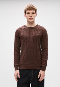 Καστόνι �πουλόβερ με καφέ πλέξη και στρογγυλή λαιμόκοψη, με ριγέ μανσέτες και στρίφωμα. Διαθέτει ένα μικρό λογότυπο στο στήθος. Φοριέται με σκούρους τζιν παντελόνια.