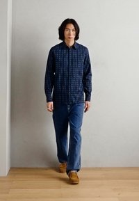 Camisa de botones azul marino con un patrón vertical, combinada con jeans azules y zapatos marrones claros, caminando sobre un suelo de madera clara contra un fondo neutro.