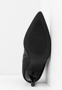 Semelle de chaussure à talon noire à bout pointu avec semelle texturée antidérapante, présentée en position verticale contre un fond blanc et un angle de mur.