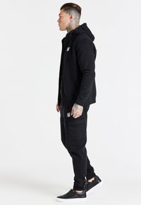 Black hoodie with front zipper and logo, paired with matching black cargo sweatpants featuring side pockets and black sneakers with white soles.