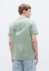 Short-sleeved, light green cotton t-shirt featuring a large gray graphic print on the back, with a soft texture and standard crew neckline.