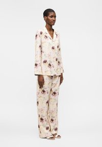 Γυναίκα που στέκεται, φοράει ανοιχτόχρωμο floral πυτζάμα σετ με μακριά μανίκια και φαρδιά παντελόνια, συνδυασμένο με λευκές ανοιχτές σανδάλια.