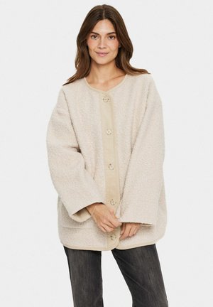 Giacca in pile beige con maniche oversize, scollo rotondo e pattina dei bottoni a contrasto. Morbida al tatto e con dettagli di trama superficiale sottili.