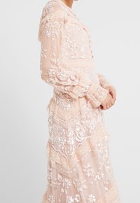 Genomskinlig persikofärgad klänning med intrikata blommönster, paljettdetaljer, spetskant, långa ärmar och en lagd kjoldesign.