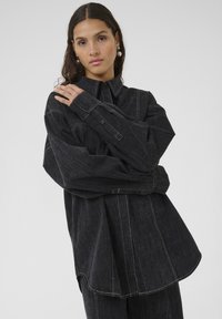 Melns džinsa krekls ar brīvu piegriezumu, ar pogu aizdari priekšā, garām piedurknēm un kontrastējošām baltām šuvēm.