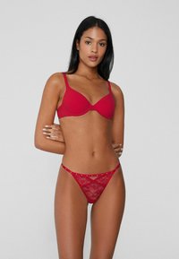 Červená souprava spodního prádla s podprsenkou s hladkou látkou a vycpávkami a průsvitnými krajkovými tangy zdobenými malými dekorativními nýty.
