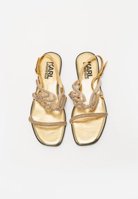 Gold metallic sandals with intricate beaded straps, featuring a smooth upper material, flat sole, and an open-toe design.